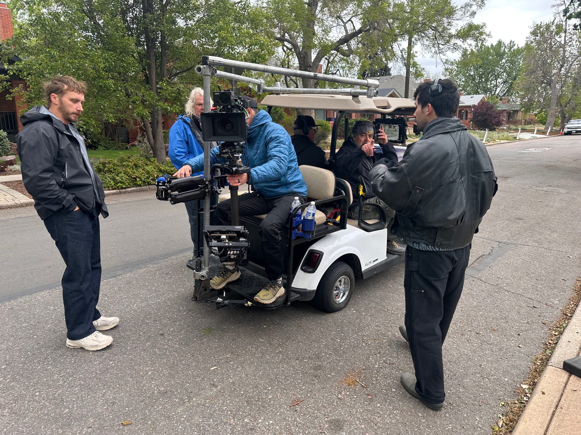 Professional cinema camera on a rear-mounted stabilizer attached to a custom pipe rig on the grip cart, with crew operating and monitoring during setup on a residential neighborhood street.